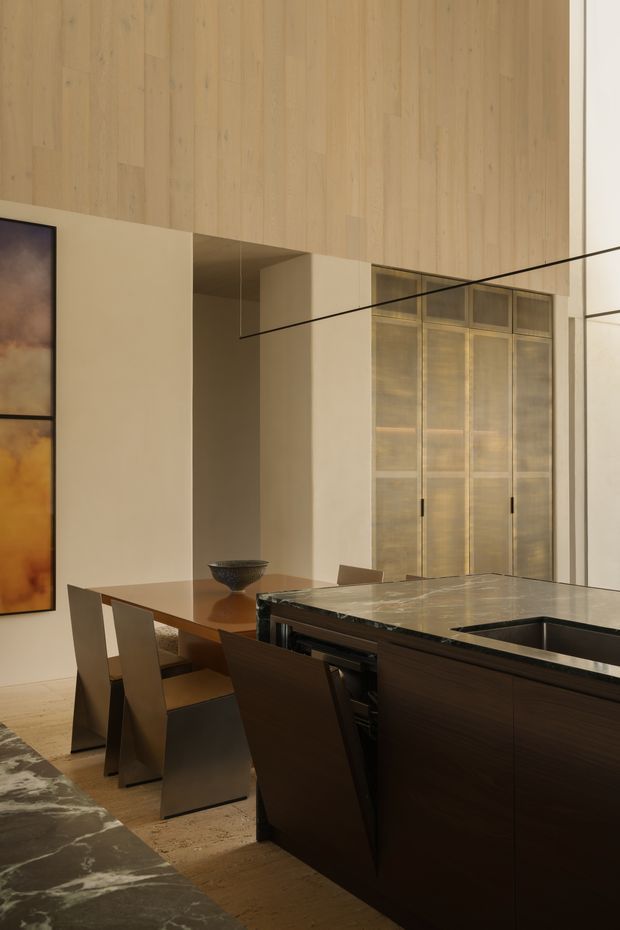 A sophisticated open-plan kitchen and dining area with a dark timber kitchen island, polished stone benchtop, integrated sink, and dark contemporary cabinetry. A warm timber-panelled double-height wall rises above the dining zone, with tall translucent glazed doors or screens providing filtered light. The room has a restrained neutral palette, sculptural dining chairs, large-scale artwork, and dramatic linear lighting.