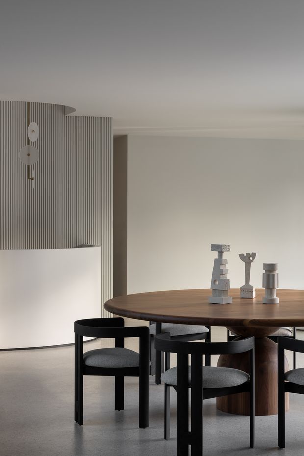 A refined contemporary dining area with a large round dark timber dining table and sculptural central pedestal, surrounded by black-framed upholstered dining chairs. The room features a pale neutral palette, polished grey concrete flooring, a curved white wall, and a vertically ribbed grey feature wall with minimalist pendant lighting and sculptural table objects.
