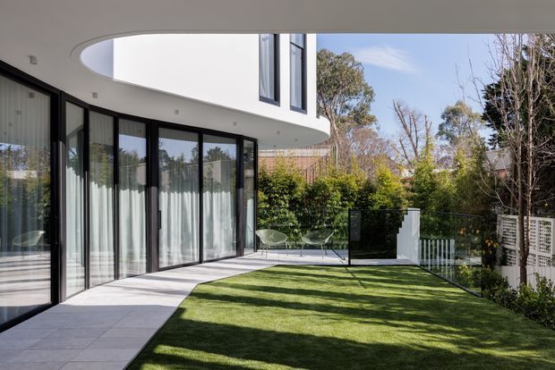 A modern residential courtyard viewed from the garden, featuring a curved white upper-level facade, extensive black-framed floor-to-ceiling glazing, a pale tiled terrace, and a neatly landscaped lawn enclosed by fencing and greenery.
