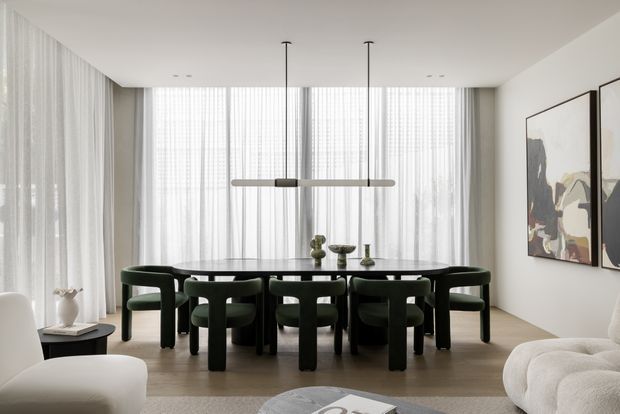 A refined contemporary dining area viewed from an adjacent living space. A large dark oval dining table is surrounded by sculptural dark green upholstered chairs, beneath a slim linear pendant light. Sheer floor-to-ceiling curtains provide soft daylight, while abstract artwork and neutral upholstered seating complete the calm, high-contrast interior.