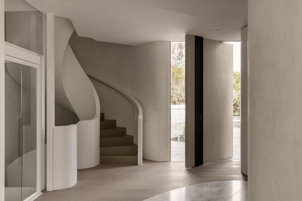 A sculptural contemporary entryway with a sweeping curved staircase, rounded plaster walls and balustrade, pale timber flooring, and a tall pivot door opening toward an outdoor area. Glazed cabinetry or a glass-panelled opening is visible at left, while the restrained neutral palette and soft daylight create a calm, gallery-like interior.