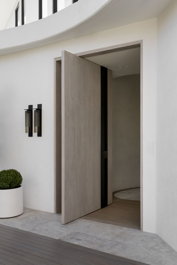 A minimalist contemporary entry viewed from an outdoor deck, featuring a tall light timber pivot door set into smooth white walls. The entry has a grey stone or concrete threshold, black and brass wall sconces, a curved overhead architectural soffit, and a white planter with a clipped green shrub. A darker interior hallway is visible through the open doorway.