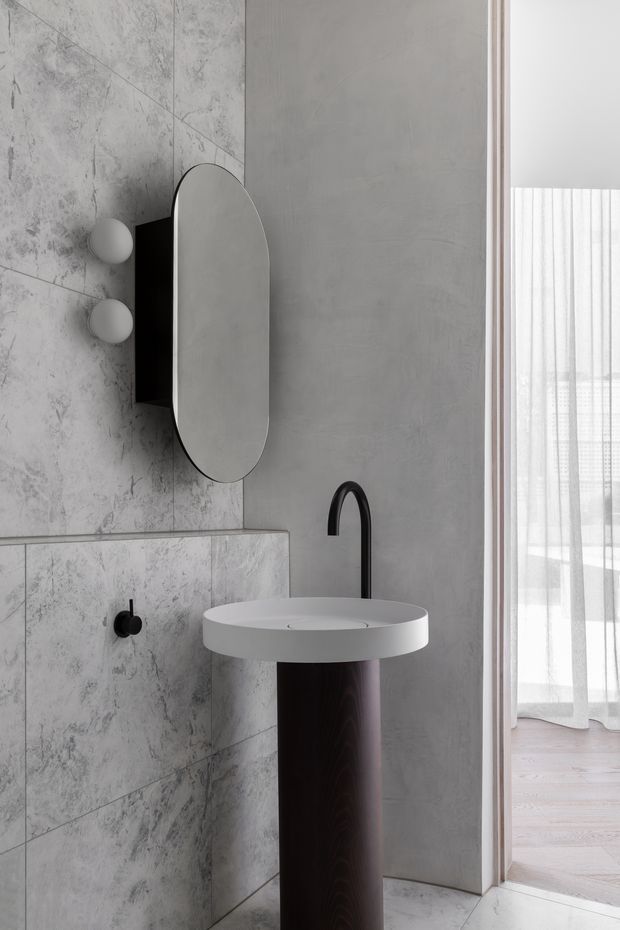 A refined contemporary bathroom vignette featuring a round white pedestal basin with a matte black curved tap, an oval wall-mounted mirror, and spherical wall lights. Large grey marble-look tiles cover the lower and left wall, while a smooth grey plaster-like wall forms the backdrop. A doorway opens to a bright adjoining room with sheer curtains and pale timber flooring.