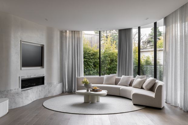 A bright, refined contemporary living room with a curved off-white sectional sofa, sculptural round coffee table, large oval rug, and a wall-mounted television above a linear fireplace set into a pale stone or plaster feature wall. Full-height sheer curtains and extensive floor-to-ceiling glazing open the room toward a leafy garden, while recessed ceiling lights and pale timber flooring complete the calm, minimal interior.