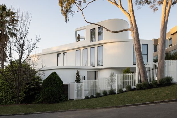 A white, multi-storey contemporary residence viewed from the street, with a sculptural curved upper-level roof or canopy, crisp rectangular glazing, smooth rendered walls, and a landscaped front garden enclosed by a white metal picket fence. Mature trees partially frame the facade.