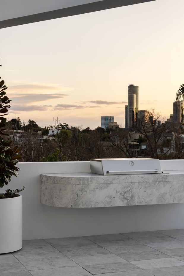 A contemporary rooftop terrace at dusk with a built-in outdoor kitchen and stainless-steel barbecue set into a projecting white marble-look counter. Large grey stone-look tiles cover the floor, while a white parapet frames a leafy suburban setting and distant high-rise city skyline. A potted plant sits at the left edge of the terrace.