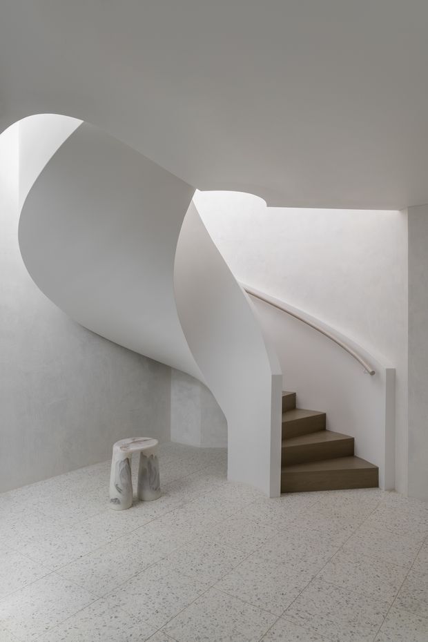A sculptural interior stairway with a sweeping, organically curved white staircase enclosure and minimalist handrail. The stairs have dark wood treads, while the surrounding walls and ceiling are smooth white plaster. The floor is light terrazzo with fine dark aggregate, and a small sculptural stool sits beside the stairs. Soft daylight and the restrained palette create a serene, gallery-like atmosphere.