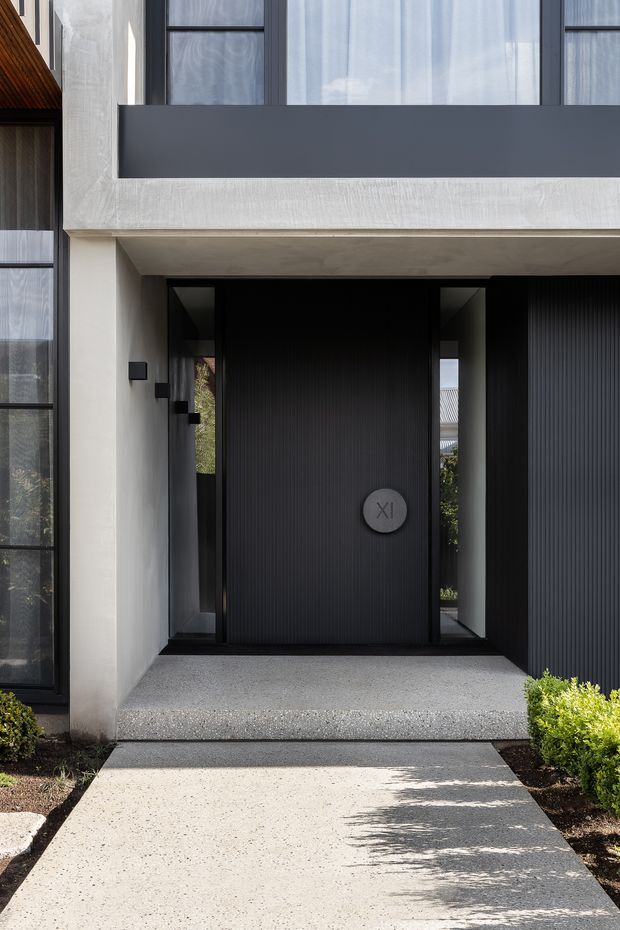 A contemporary residential entrance viewed from outside, featuring a tall black ribbed front door with narrow glazed sidelights, a recessed porch, pale textured render, charcoal window framing, and a projecting upper-level balcony or facade panel. A light exposed-aggregate pathway leads to the entry, with neatly landscaped planting along both sides.