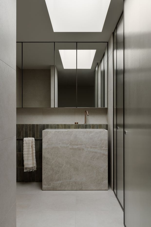 A refined contemporary bathroom featuring a monolithic rectangular stone vanity with an integrated basin, a slim brushed metal faucet, large wall mirrors, and dark vertical tiled detailing behind the basin. Full-height textured grey cabinetry or wall panels line the right side, while large rectangular ceiling skylight-style light wells create a bright geometric contrast. The palette is restrained in warm greys, stone, charcoal, and subtle bronze.