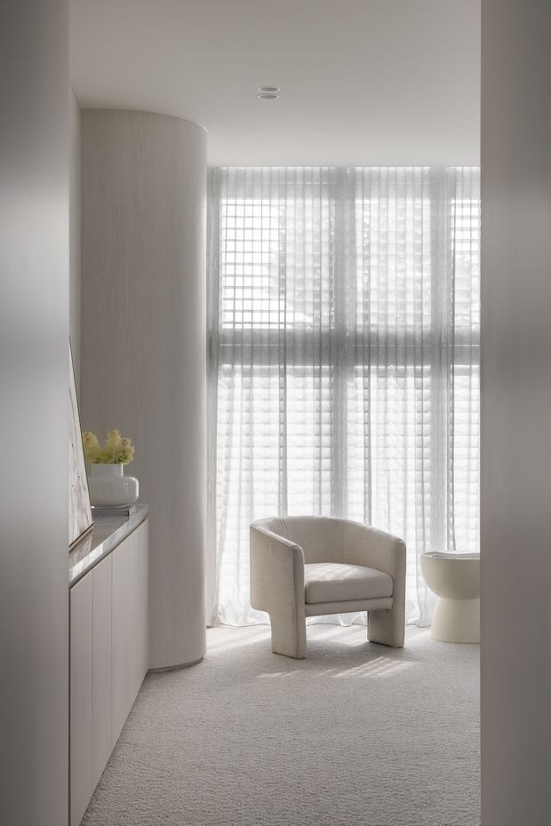 A softly lit, minimalist interior with a pale carpeted floor, curved light-wood or wood-finished partition, built-in cabinetry, and sheer floor-to-ceiling curtains over a gridded window. A rounded upholstered armchair sits beside a sculptural white pedestal table, creating a calm, monochromatic seating nook.