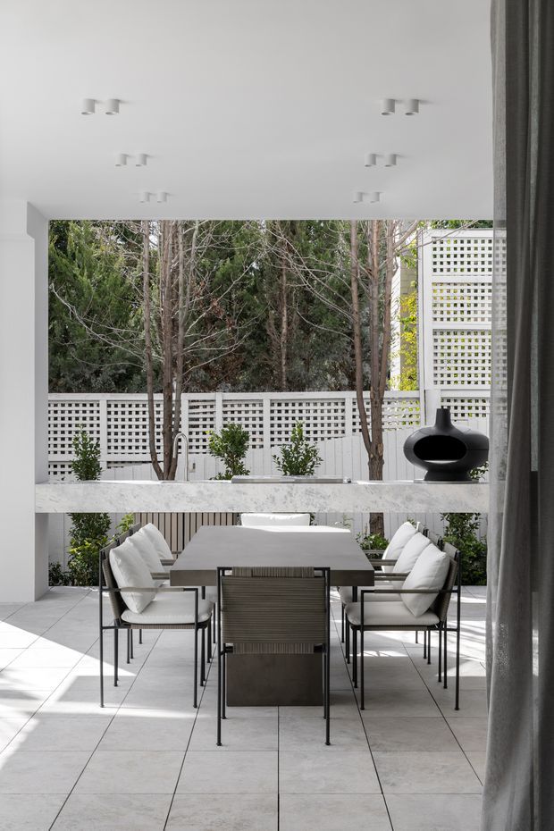 A bright, refined indoor-outdoor dining area opening onto a planted courtyard. A dark rectangular dining table is surrounded by upholstered dining chairs, with a long pale stone serving ledge behind it and a black freestanding grill or outdoor cooker. Large pale floor tiles, white walls, recessed ceiling spotlights, sheer curtains, and perforated white masonry screens create a clean contemporary setting with strong garden views.