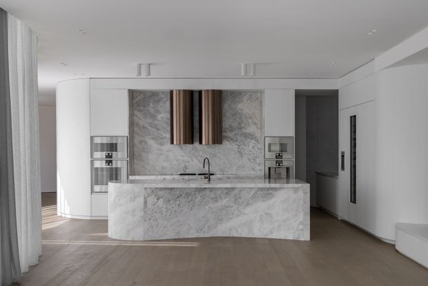 A sleek, minimalist kitchen with a large waterfall island clad in dramatic grey-veined stone, matching stone splashback, white integrated cabinetry, and dark timber vertical accents. Built-in ovens and appliances are set into the cabinetry, with a concealed or partially visible pantry area at right. The kitchen opens to a light-filled living area with full-height sheer curtains and warm timber flooring.