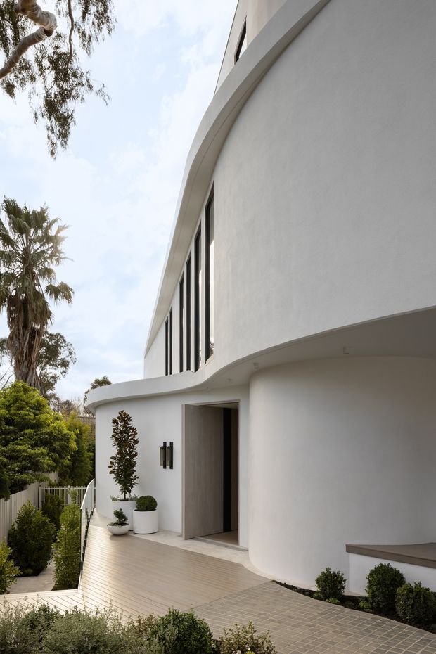 A refined white-rendered residential exterior with strong curved walls, rounded roofline edges and tall narrow black-framed windows. The entrance opens onto a pale timber-toned deck and a grey tiled pathway, with clipped shrubs, potted plants and mature palm and eucalyptus trees framing the approach. The composition highlights smooth sculptural forms and minimalist detailing.