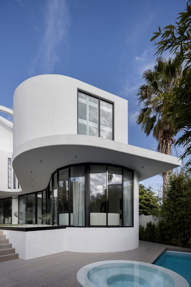 A striking contemporary two-storey residential exterior with smooth white curved walls, a broad projecting curved canopy, and extensive black-framed glazing. The building opens onto a timber deck with a small pool and circular spa in the foreground, while tropical palms and dense planting surround the property. The image is photographed from outdoors under a clear blue sky.