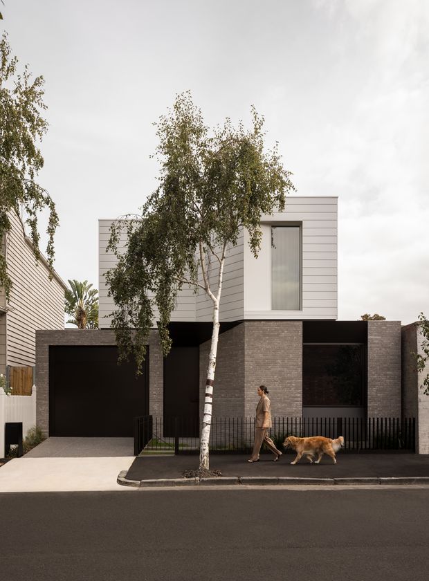 Street-facing view of a contemporary multi-level urban residence with a rectilinear composition. The lower level is clad in dark textured brick and includes a black garage door, recessed entry, and large dark window openings. The upper level has pale horizontal cladding, a tall narrow window, and a flat roof. Black metal fencing, a small front garden, paving, and a birch tree line the footpath.