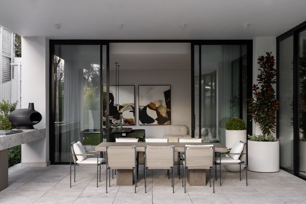 Contemporary covered outdoor dining terrace adjoining a glazed living room. The space features a long dining table with multiple woven dining chairs, pale tiled flooring, black-framed floor-to-ceiling glazing, white walls and ceiling, large cylindrical planters, and a built-in stone-topped counter with sculptural black decor. Abstract artwork and a sofa are visible inside through the glass doors.