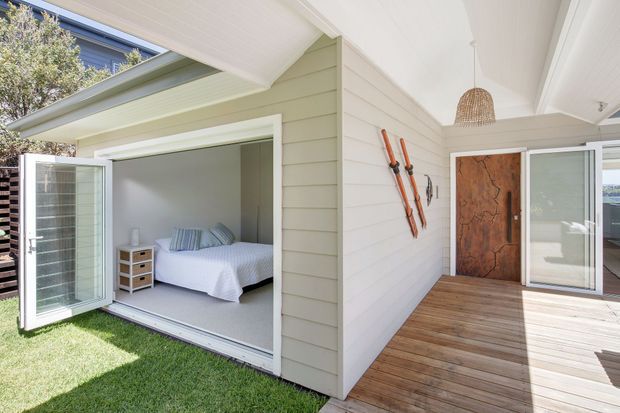 Bright coastal-style residential exterior showing a compact bedroom pavilion opening onto a timber deck. The pavilion has pale horizontal weatherboard cladding, a white pitched ceiling and trim, large glazed sliding and folding doors, and a rustic timber entry door. A bed and bedside storage are visible inside, while the deck connects to a lawn and the main house.