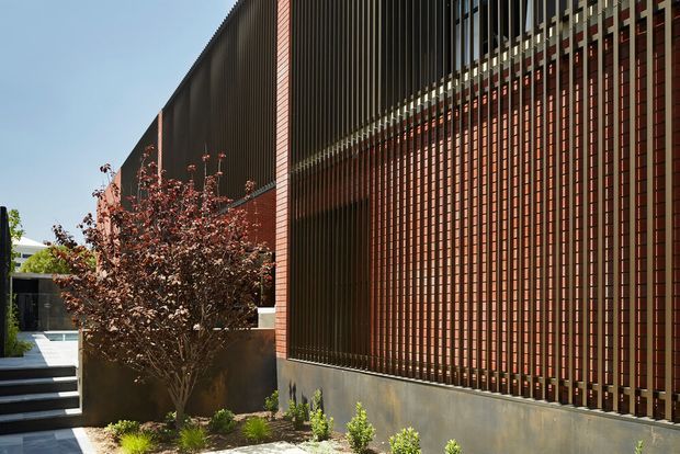 Exterior architectural view of a contemporary building featuring a long red-brick wall screened by closely spaced vertical dark metal fins, with a contrasting concrete base and landscaped entry walkway. Strong linear rhythm, deep shading, and a modern institutional or commercial facade are prominent.