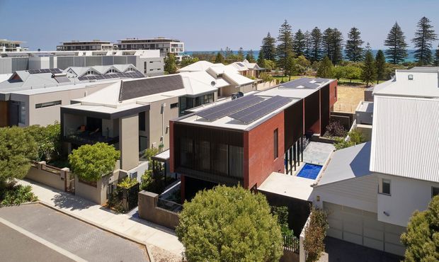 Elevated view across a contemporary coastal residential neighbourhood, showing several modern homes with flat and skillion roofs, rooftop solar panels, dark and light cladding, landscaped gardens, and a visible swimming pool. Tall coastal trees and blue water appear in the background.