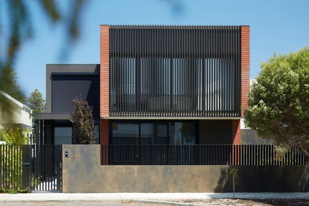 Street-facing view of a contemporary two-storey residence with a strong rectilinear composition. The facade combines dark rendered wall panels, red brick vertical piers, extensive upper-level black vertical screening, dark metal fencing, and large glazed openings. A rendered boundary wall and landscaped planting frame the property.