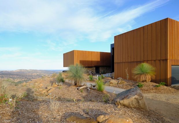Exterior view of a contemporary, angular building composed of warm vertical timber-clad volumes set into a rocky, arid landscape. Large glazed openings and dark recessed areas contrast with the timber facade, while drought-tolerant grasses, scattered boulders and gravel create an integrated desert-style landscape. Expansive valley and mountain terrain are visible beyond the property under a bright blue sky.
