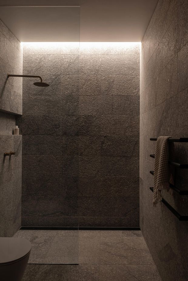 A compact, dark-toned bathroom with a walk-in shower enclosed by clear glass. Large textured grey stone-look tiles cover the walls and floor, with a concealed linear drain, matte black shower fittings, and a black heated towel rail holding a textured towel. Subtle concealed lighting runs along the upper shower wall, creating a moody and refined atmosphere.