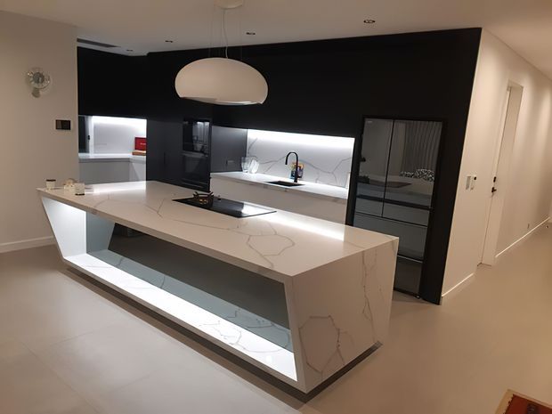Contemporary kitchen featuring a large white veined stone island with an illuminated open display base, integrated cooktop, black cabinetry and backsplash, under-cabinet lighting, and a sculptural pendant light. A hallway and additional built-in cabinetry are visible beyond the kitchen.