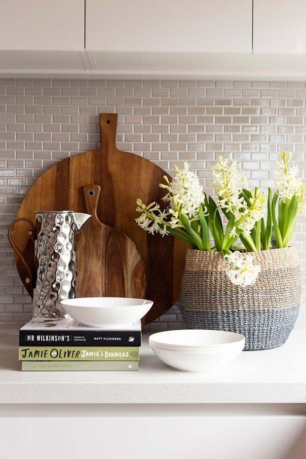 A styled kitchen vignette featuring a pale pink-grey brick-pattern tile splashback, white cabinetry and a light benchtop. Wooden cutting boards lean against the splashback beside a hammered silver metal jug, white ceramic bowls, stacked cookbooks and a large woven planter filled with white flowers and green foliage.