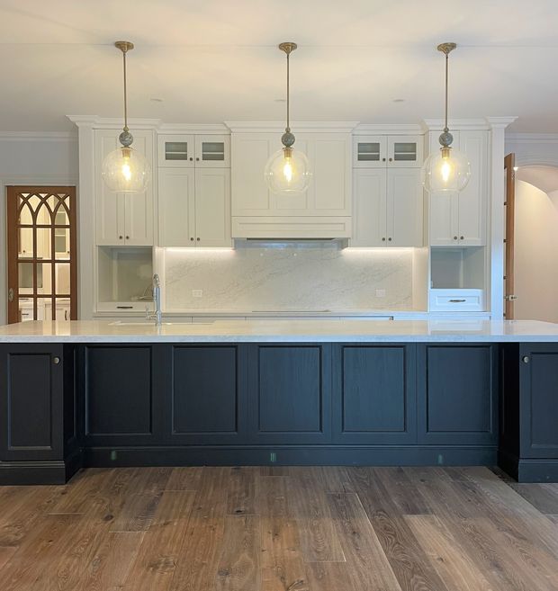 A spacious, traditional-style kitchen centred on a large island. The room features dark navy-blue raised-panel island cabinetry, white shaker-style wall cabinetry, a pale stone benchtop and illuminated stone splashback, warm timber flooring, brass-toned hardware, and three clear-glass pendant lights. Arched timber-framed glazing and decorative mouldings reinforce the classic architectural character.