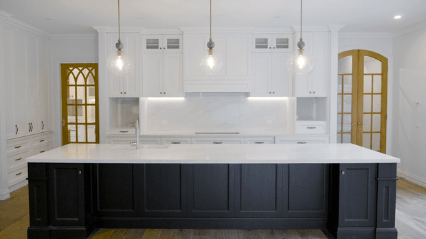 Bright, spacious kitchen with a large white island featuring dark charcoal paneled cabinetry, white shaker-style perimeter cabinetry, a pale stone benchtop and backsplash, arched timber-framed glazed doors, and three glass pendant lights. The room has a refined traditional architectural character with strong black-and-white contrast and warm timber flooring.