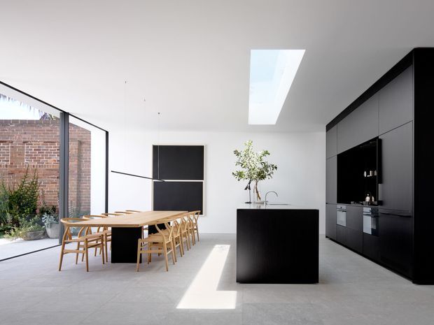 Bright contemporary kitchen and dining area with a large black island, full-height matte dark cabinetry, pale stone flooring, timber dining table and chairs, expansive glazing, and a linear skylight. A brick wall and planted courtyard are visible outside.