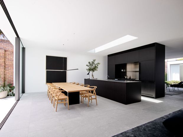 A bright, open-plan contemporary kitchen and dining area with a long light-wood dining table and matching chairs beside a large black kitchen island and full-height dark cabinetry. White walls and ceiling, pale large-format flooring, a linear skylight, expansive glazing, and exposed brick visible at the edge create a clean, modern space.