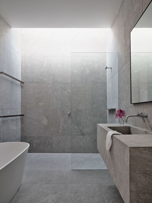 Contemporary minimalist bathroom featuring large-format grey stone-look wall and floor tiles, a frameless glass walk-in shower, freestanding white bathtub, floating stone vanity with integrated basin, wall-mounted chrome tapware, and a large mirror. The space is calm, refined, and predominantly monochromatic with subtle natural texture.