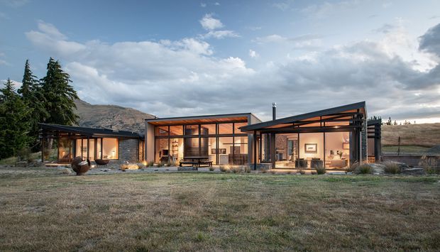 Wide exterior view of a low, modern rural residence arranged as several connected pavilion-like volumes. The house has extensive floor-to-ceiling glazing, dark metal structural framing and cladding, warm timber-toned interior illumination, and largely flat or very low-pitched roof forms. A broad grassed foreground, mature conifers, and rugged hills form the surrounding setting.