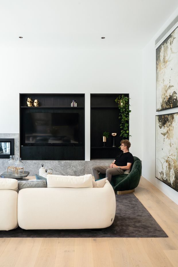Bright contemporary living room with a large cream sectional sofa, dark green lounge chair, black built-in television and display cabinetry, grey stone feature wall, pale timber flooring, and large abstract artworks. Recessed ceiling lights and concealed linear air-conditioning vents contribute to a clean, refined interior.