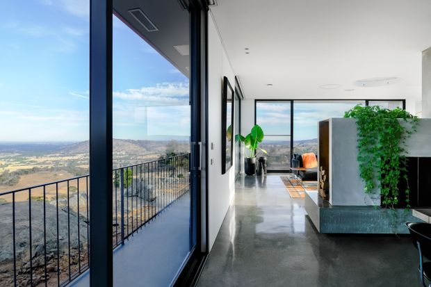 A bright contemporary interior opening onto a balcony or terrace with expansive views over a dry rolling landscape. The long open-plan space has polished concrete flooring, white walls, black-framed floor-to-ceiling glazing, a dark kitchen or joinery volume, indoor greenery, and a glimpse of an orange lounge chair. A black metal balustrade runs along the exterior edge.