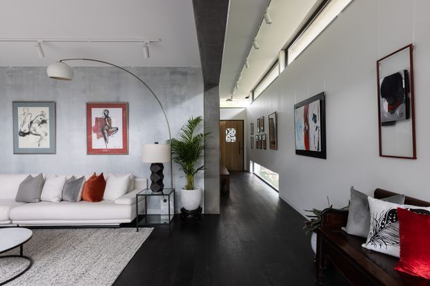 A refined contemporary living room and adjoining hallway with dark timber flooring, white walls, and a textured concrete feature wall. The space includes a white sofa with neutral and red cushions, a sculptural floor lamp, potted plant, framed artwork, a glass side table, and a dark timber console. Linear ceiling lighting and clerestory glazing create a bright, gallery-like interior.