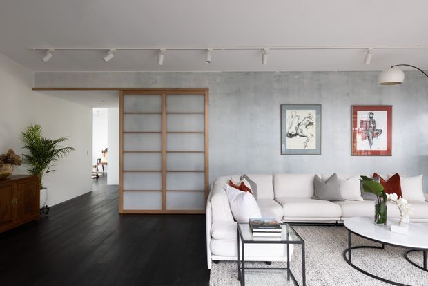 A bright, contemporary living room with a large white sectional sofa, round white coffee table, black-framed side table, and patterned rug. The room features dark timber flooring, a textured grey concrete-look wall, a timber-framed frosted glass sliding partition, track lighting, and modern artwork. A hallway and part of another room are visible beyond the partition.