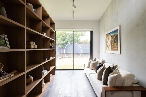 A compact, light-filled living room with a long low sofa, neutral cushions, a full-height timber built-in shelving wall, pale timber flooring, and a large black-framed sliding glass door opening to an outdoor area. A framed artwork and minimalist track lighting complete the restrained contemporary interior.