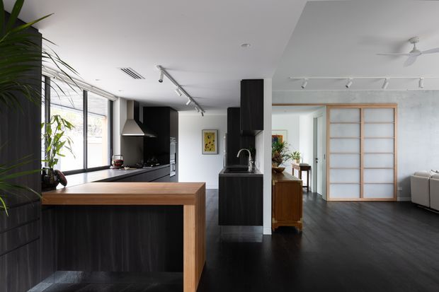 Open-plan contemporary kitchen and living area with dark timber cabinetry, a warm timber island surround, black flooring, white walls and ceiling, and large glazed windows. Track lighting, minimalist fittings, and a timber-framed sliding screen define the space, with a living room visible beyond the kitchen.