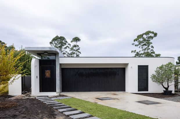 Contemporary single-storey residential facade with a flat roof, smooth white rendered walls, a dramatic black ribbed garage door, and a recessed black entry door framed by a raw concrete portal. A stepping-stone path crosses a small lawn toward the entrance, with minimalist landscaping and mature trees surrounding the property.
