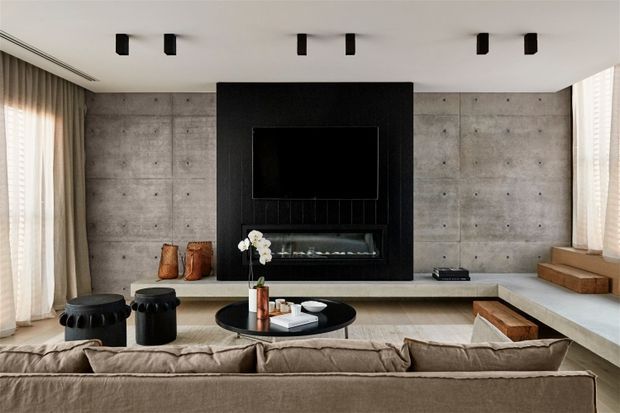 Contemporary living room with a large concrete feature wall, a black built-in media and fireplace surround, integrated low bench seating, floor-to-ceiling curtains, and a neutral sectional sofa arranged around a round coffee table. Minimal black ceiling spotlights and restrained furnishings create a refined, modern atmosphere.