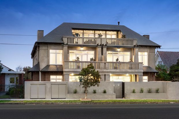 A symmetrical multi-storey residential building photographed at dusk from across the street. The facade combines dark brickwork, rendered concrete or stucco surfaces, large illuminated windows, and two levels of projecting balconies with vertical timber or painted balustrades. A broad hipped roof with dark shingles and dormer-like upper glazing tops the building, while a low rendered front boundary wall and planted verge sit along the street.