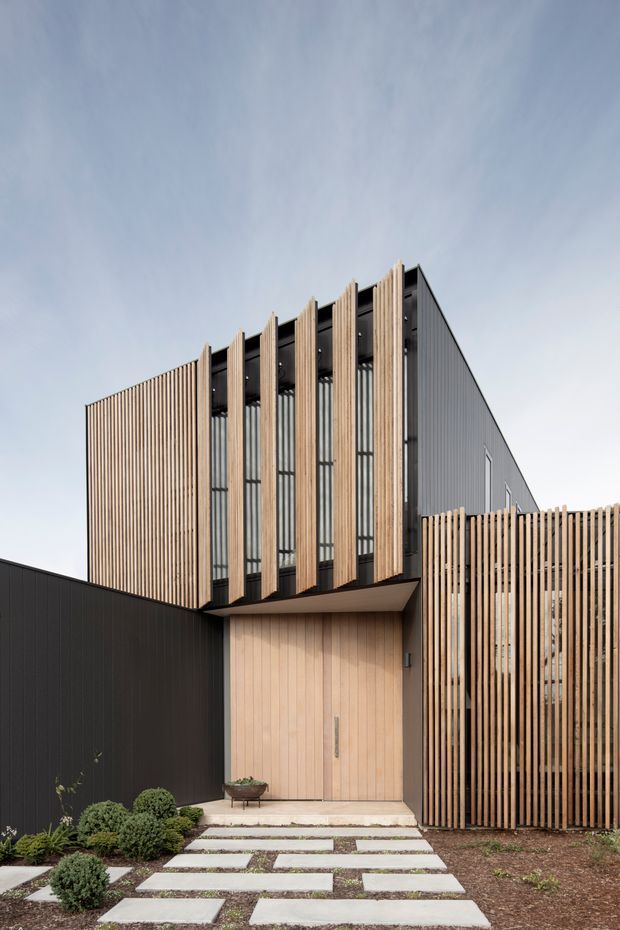 Exterior architectural photograph of a contemporary multi-level building entrance. The facade combines dark vertical metal cladding with extensive vertical timber battens and tall glazed openings. A recessed timber-clad doorway sits beneath a projecting canopy, with a timber slat privacy screen extending along the right side. The landscaped approach uses large rectangular stepping slabs, gravel or soil, and clipped shrubs.