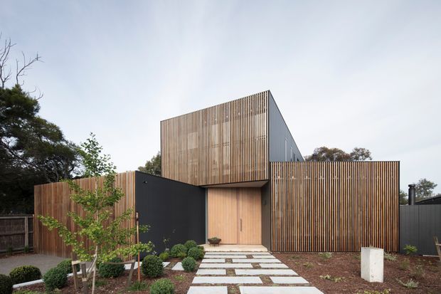 Contemporary residential facade composed of vertical timber screening, dark painted wall panels, and a recessed timber entry door. The two-storey geometric form is set behind a landscaped front garden with clipped shrubs and a stepping-stone path, creating a clean, privacy-focused street elevation.