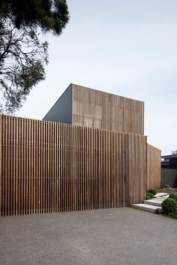 Street-level exterior view of a contemporary multi-volume building screened by closely spaced vertical timber battens. A tall upper volume with a steeply sloping roof or wall plane rises behind the timber-clad frontage, with a paved driveway and a landscaped stepping-stone path leading toward the entrance. The composition is minimalist and strongly geometric, with dark metal and timber elements.
