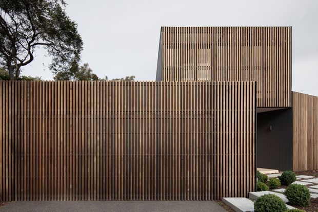 Exterior architectural view of a contemporary multi-level building screened by closely spaced vertical timber battens. The facade forms layered volumes with a tall upper section, recessed dark entry or opening, and a landscaped foreground with geometric stepping stones and clipped shrubs. Mature trees are visible in the background.