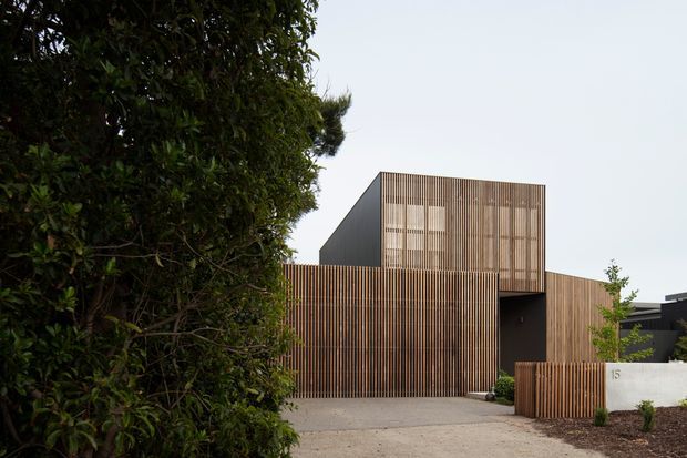 Exterior view of a contemporary multi-level residence with strong geometric massing and extensive vertical timber screening. A tall upper volume sits above a lower timber-clad frontage, with a dark recessed entry and a broad paved driveway. Dense hedging and simple landscaped planting frame the building.