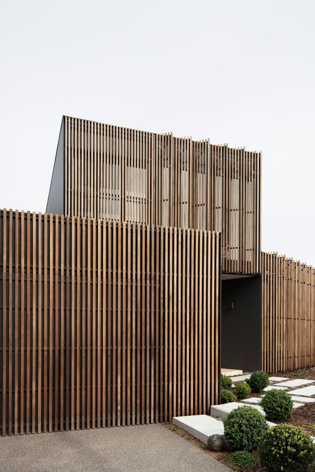 A contemporary multi-level building facade enclosed by closely spaced vertical timber battens. The slatted screen creates layered volumes, filtered openings, and a recessed dark entry. A minimalist landscaped path with rectangular stepping stones and clipped shrubs leads toward the entrance.