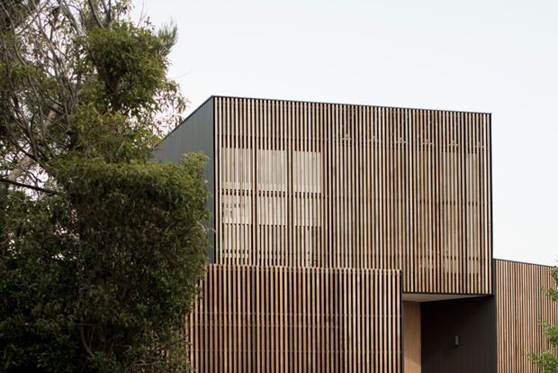 Exterior view of a contemporary, box-like building screened by closely spaced vertical timber slats. The slatted facade conceals upper-level glazing and wraps around a recessed dark entry, with mature landscaping partially obscuring the lower left. The composition is minimalist and rectilinear, with muted natural timber tones against a dark wall plane.
